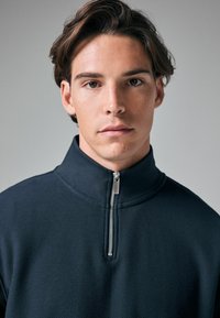 Marineblauer Quarter-Zip-Pullover aus strukturiertem Stoff. Verfügt über einen Stehkragen und einen metallenen Reißverschluss, mit einer glatten Oberflächenverarbeitung am Halsausschnitt.