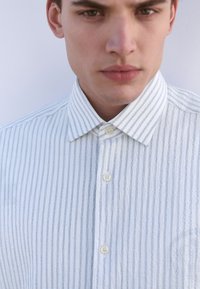 Chemise à rayures bleu clair et blanc avec fermeture à boutons, col classique, texture subtile et boutons en plastique.