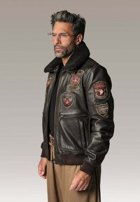 Homme avec des cheveux grisonnants et des lunettes, portant une veste bombardier marron avec de multiples écussons de style militaire et un pantalon beige, se tenant de côté.