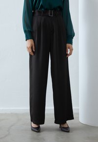 Pantalons noirs à jambes larges avec plis, taille haute et ceinture. Tissu lisse et coupe ajustée, associés à des escarpins noirs pointus.