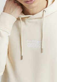 Felpa beige con cappuccio e cordino, caratterizzata da un testo bianco ricamato "SLD-EST.1999" e un motivo a righe. Tessuto morbido e design minimalista.