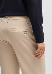Pantaloni beige con una texture liscia e una tasca posteriore con il logo "BOSS" nero. Indossati con una camicia a maniche lunghe scura.