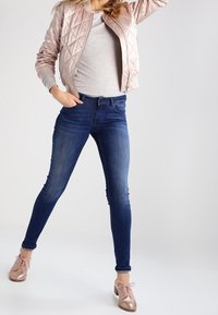 Vadderad ljusrosa bomberjacka, grå t-shirt, mörkblå skinny-jeans och glänsande roséguldsskor, poserad mot en neutral bakgrund.