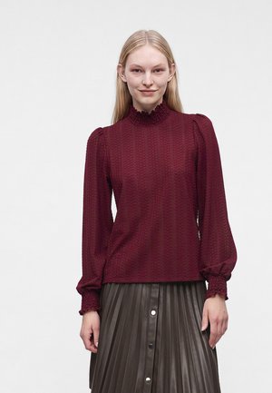 Jeune femme portant un haut bordeaux texturé à manches longues avec un col haut à volants et une jupe plissée foncée avec des boutons sur le devant.