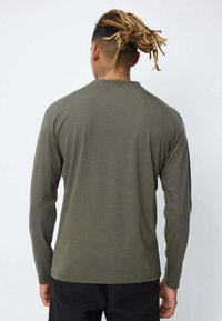 Homme avec des dreadlocks attachées, portant une chemise à manches longues olive et un pantalon noir, tourné le dos contre un fond léger uni.