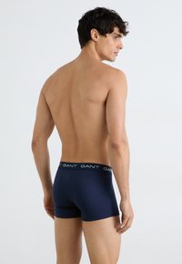Marineblaue Boxer-Shorts mit einer weichen Textur und einem kontrastierenden schwarzen Taillenbund mit wiederholt eingearbeitetem weißen Branding. Modell zeigt die Rückansicht.