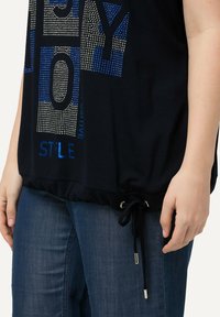Černé krátké tričko s modro-bílým nápisem z rhinestone, nařaseným lemem s nastavitelnou šňůrkou, nošeno s modrými džíny.