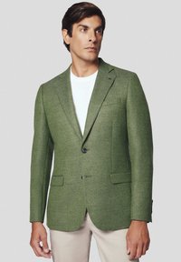 Zielony żakiet wykonany z teksturowanej tkaniny, z pojedynczym guzikiem, klapami w szpic i dwoma przednimi kieszeniami. Noszony na białej koszuli.