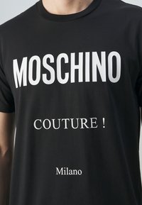 Svart T-shirt med vit text som läser "MOSCHINO" i stora bokstäver, "COUTURE!" nedanför, och "Milano" i bottencentret.