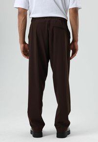 Bruine, loszittende broek met een elastische tailleband en een enkele achterzak, gemaakt van gladde stof, gecombineerd met zwarte schoenen.