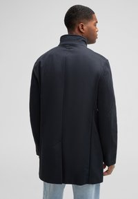 Navy long coat with high collar, smooth texture, single back vent, and minimalistic design. Features discreet logo on collar.