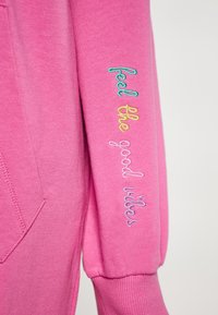 Felpa rosa con tasca a marsupio; presenta ricami colorati sulle maniche con la scritta "senti le buone vibrazioni". Materiale morbido con polsini a coste.
