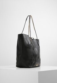 Mustassa nahkaisessa kassi, jossa on kohokuvioitu kukkakuviot, kaksoiskahvat ja metallivetoketjuyksityiskohta. Rakenteeltaan teksturoitu ja tilava muotoilu.