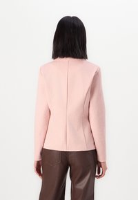 Svetle růžová fitted bunda s švem na zadní straně a vysokým límcem, kombinovaná s hnědými kalhotami z umělé kůže. Hladká textura.