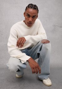 Suéter de canalé crema, jeans rasgados de color azul claro y zapatillas blancas. Modelo agachado contra un fondo gris, mostrando el ajuste relajado y los detalles.