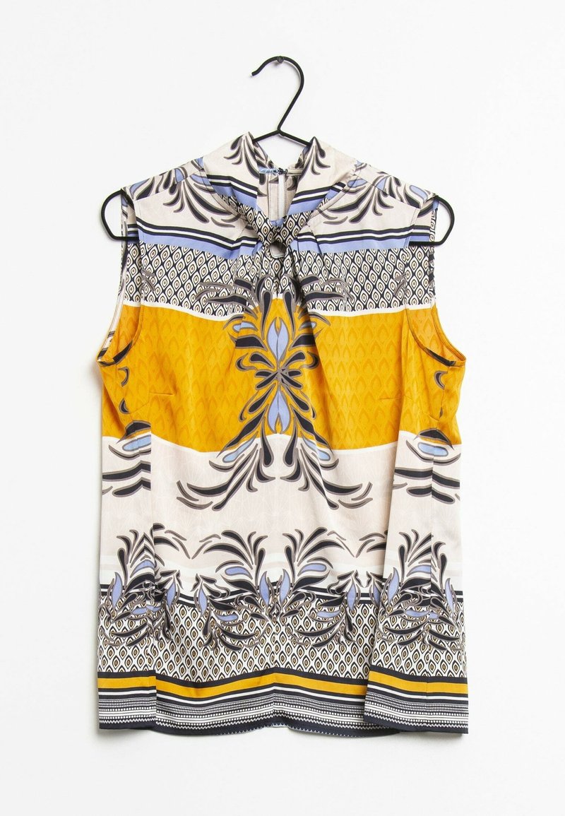 Blouse sans manches avec des motifs ornementaux et géométriques jaunes, blancs, noirs et bleus, suspendue sur un cintre noir sur fond blanc.