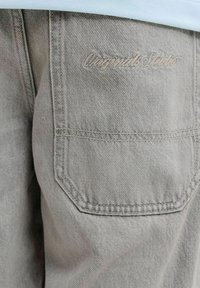 Κοντινή λήψη της πίσω τσέπης γκρι τζιν με διακριτικό κεντημένο λογότυπο "Olympia Sports" πάνω από τη ραφή της τσέπης.