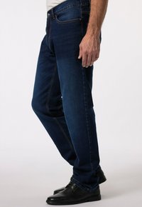 Tmavě modré džíny se rovný střih, s kontrastním prošíváním, pěti kapsami a zapínáním na knoflík. K tomu jsou obuty černé boty.