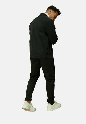 Veste noire avec un col, associée à un pantalon cargo noir. Le mannequin porte des baskets blanches et est positionné avec un léger tourné.