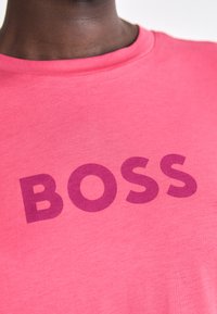 Pink cotton t-shirt with short sleeves, featuring bold purple text spelling "BOSS" across the front. Smooth texture, classic crew neck design.