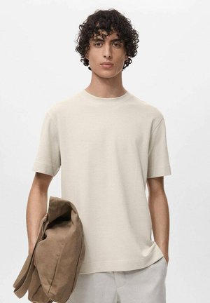 Cremefarbenes, kurzärmeliges Strick-T-Shirt mit Rundhalsausschnitt, getragen mit einer braunen Stofftasche, die über dem Arm hängt. Glatte Textur und lockere Passform.