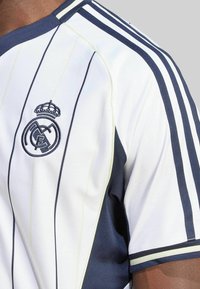 Camiseta de fútbol blanca con acentos en azul marino, presenta rayas verticales, cuello redondo y diseño de escudo bordado en el lado izquierdo del pecho.