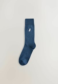 Tamsiai mėlyna kojinė su raukšlėtu viršumi ir tekstūruotu kūnu. Šone yra maža išsiuvinėta balta katytės dizaino detalė.