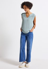Lichtblauw, getextureerd mouwloos topje met een V-hals en decoratieve stiksels, gecombineerd met oversized blauwe jeans en lichtgekleurde sneakers.