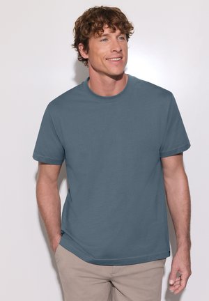 Homme aux cheveux bruns bouclés portant un t-shirt bleu à manches courtes et un pantalon beige, souriant et regardant légèrement sur le côté.
