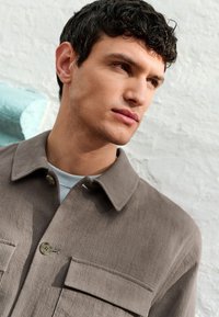 Camisa con cuello en tejido texturizado marrón claro, con dos bolsillos en el pecho y cierre de botones, superpuesta sobre una camisa azul claro.