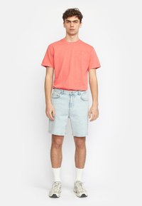 Koraalkleurig t-shirt met korte mouwen gecombineerd met lichtblauwe denim shorts. Model draagt witte sokken en grijze sneakers. Eenvoudig ontwerp, geen zichtbare patronen.