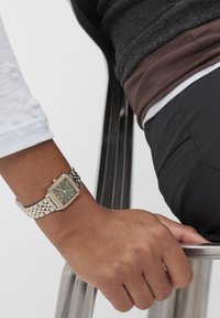 Silber- und goldfarbener quadratischer Uhr mit einem dunkelgrünen Zifferblatt und goldenen Stundenmarkierungen, getragen am Handgelenk, mit einer Hand, die auf einem Metallstuhl ruht.