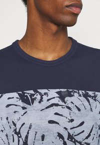 Granatowa koszulka z okrągłym dekoltem, z kontrastowym jasnoszarym wzorem liści na przodzie, wykonana z miękkiej, teksturowanej bawełny.