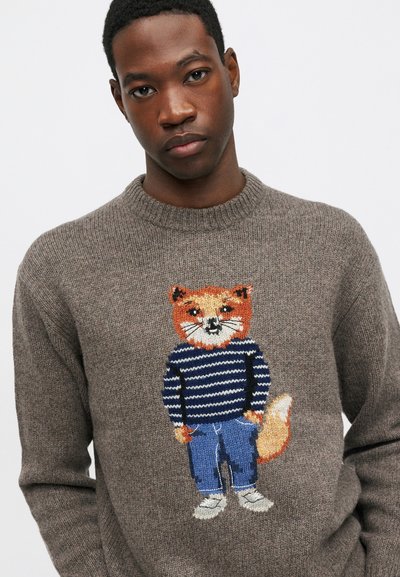 Jauns vīrietis, tērpies brūnā adītā džemperī ar karikatūras lapsas attēlu, valkā strikti svītrainus kreklus, zilos bikses un baltas kurpes, rokas ieliktas kabatās.