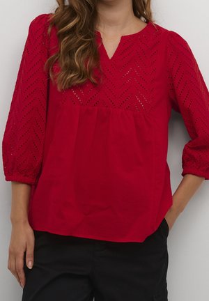 Vrouw draagt een rode blouse met lange mouwen met oogjes-chevronpatroon bovenaan en zwarte broek, staand met één hand in de zak.