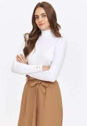 Biała longsleeve koszulka z golfem o gładkiej fakturze, z złotymi guzikami przy mankietach, w połączeniu z brązowymi spodniami o wysokim stanie i tapered fit.