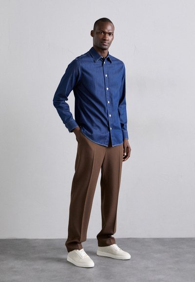 Camicia in denim azzurro scuro con bottoni, abbinata a pantaloni marroni su misura e sneakers bianche. Texture liscia, vestibilità standard e bottoni a contrasto.