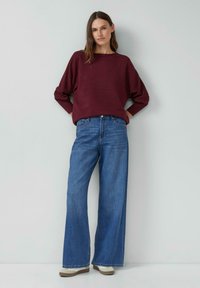 Bordowy sweter w paski z szerokimi rękawami, połączony z wysokim stanem, luźnymi niebieskimi jeansami i jasnymi butami.