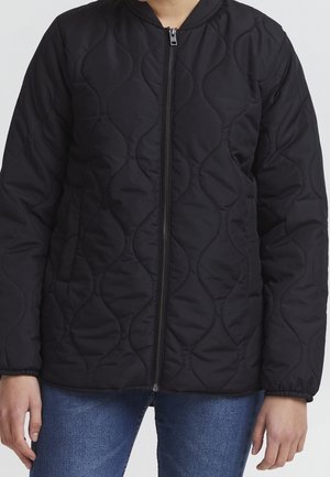 Quilted black zip-up jacket worn with blue jeans, featuring a subtle wavy stitch pattern and elastic cuffs on the sleeves.
