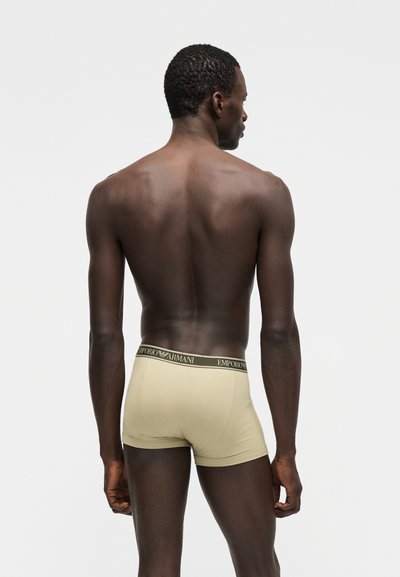 Άνδρας που στέκεται με την πλάτη στραμμένη, φορώντας μπεζ boxer Emporio Armani με λάστιχο μέσης με λογότυπο, σε απλό φωτεινό φόντο.
