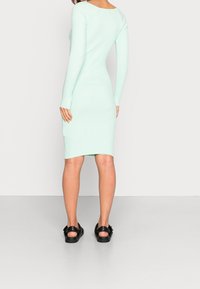 Vestido de punto acanalado verde menta con mangas largas y silueta ajustada, que presenta un cuello redondo y un dobladillo a la altura de las rodillas. Combinado con sandalias negras.