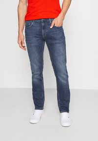 Jeans oscuros de hombre, cortados slim, con un ligero desgaste. Cuentan con cinco bolsillos, trabillas para el cinturón y cierre de botón. Combinados con zapatillas blancas.