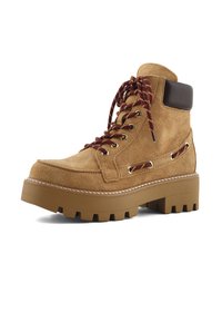 Botín de ante color canela con acentos de cuero marrón, cordones marrones y amarillos, suela de goma gruesa y detalles de costura visibles.