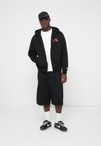 Sweatshirt com capuz preto com logótipo rosa, t-shirt branca, calções pretos até ao joelho, meias brancas e ténis pretos com detalhes brancos.