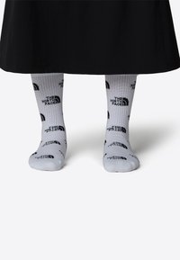 Vita strumpor med svart varumärke, prydda med upprepade logotyper av "The North Face." Ribbad textur, synlig ovanför nederkanten på den svarta klädesdelen.