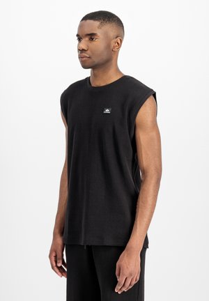 Junger Mann trägt ein ärmelloses schwarzes strukturiertes Hemd mit einem kleinen Logoaufnäher auf der Brust, steht vor einem weißen Hintergrund.