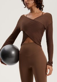 Barna bordázott, hosszú ujjú felső, keresztezett dizájnnal, melyet magas derekú barna leggings-szel visel. A modell egy fém színű fitneszlabdát tart a kezében.
