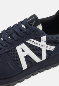 Baskets de sport bleu marine en matériaux mesh et daim, avec une marque blanche, des lacets plats et une semelle noire texturée.