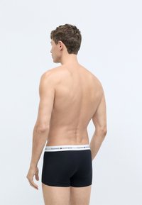 Sorte boxerbriefs med en hvid linning, der viser "TOMMY HILFIGER" logoet, lavet af et glat stof, vist fra bagsiden.