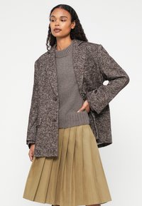 Pruun herringbone-mustriga bleiser, millel on vööri-särk ja hall kudumisega sviiter ning khaki voltidega seelik. Tekstuuriga kangaste detailid on nähtavad.